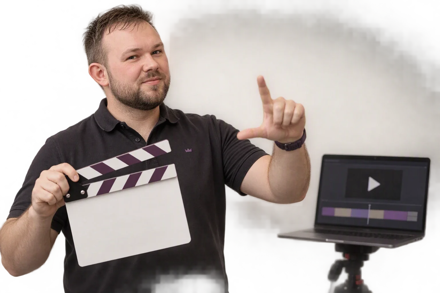 Ein Mann hält eine Filmklappe mit aubergine-weißen Streifen und formt mit der anderen Hand einen Bildausschnitt; hinter ihm zeigt ein Laptop einen Videoschnitt mit Abspielknopf und Zeitleiste.